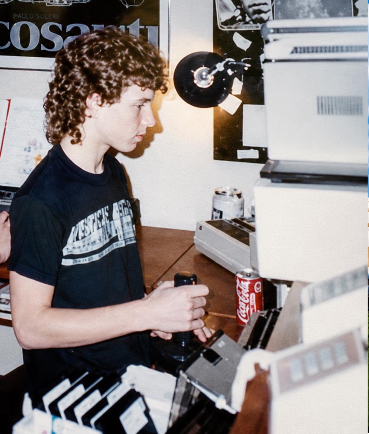 A teenager with curly hair sits at a Commodore computer setup surrounded by floppy disks, a joystick, disk drives and posters
